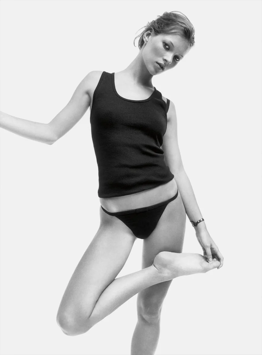 Kate Moss in a black vest and underwear against a plain backdrop, photographed by David Sims for Calvin Klein in 1993