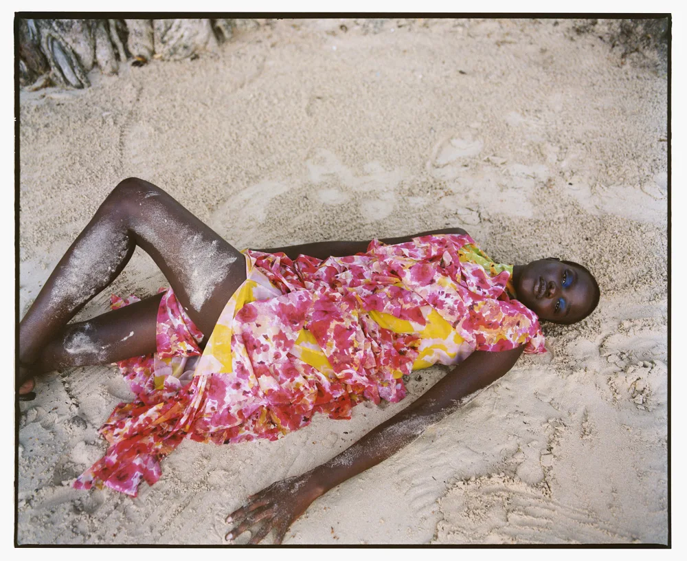 Model reclining in a bright floral dress against textured pale stone, photographed by Nadine Ijewere